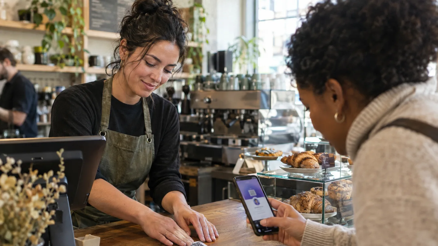 A cafe owner sticking a QR code label onto the counter by the register while a customer points a phone camera at it